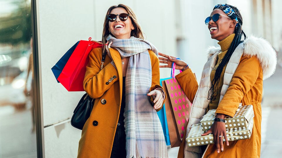 two women shoppers talking and laughing while carrying shopping bags outside a retail store