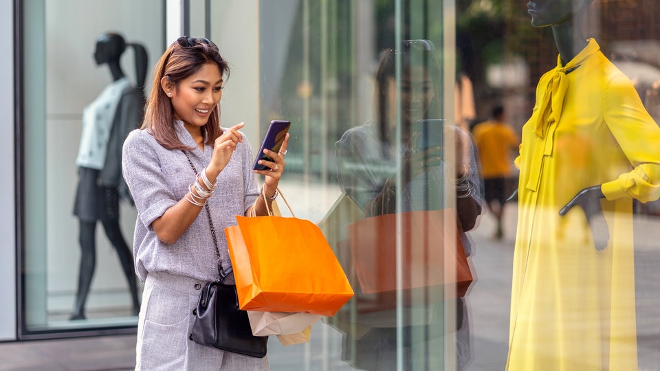 woman shopper carrying shopping bags using her phone to find information on the item displayed in the retail apparel store window